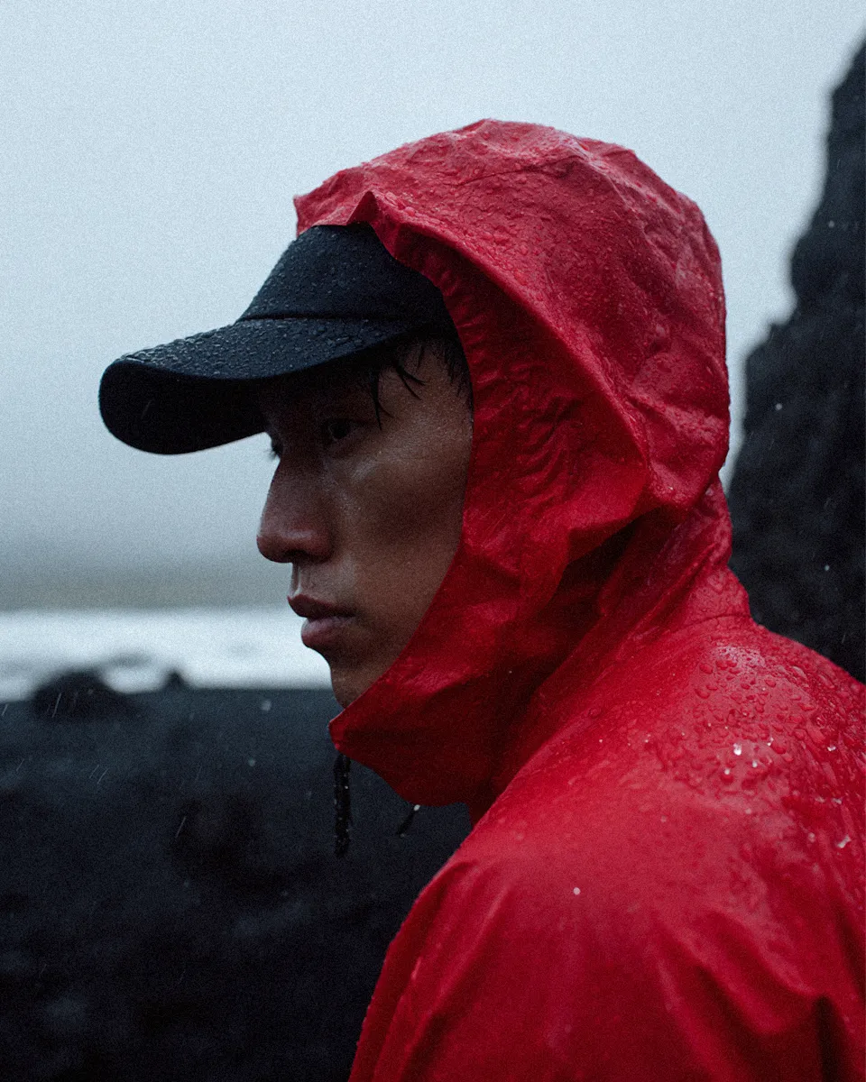 Person in a red rain jacket and black cap looking serious on a rainy day near dark rocks.