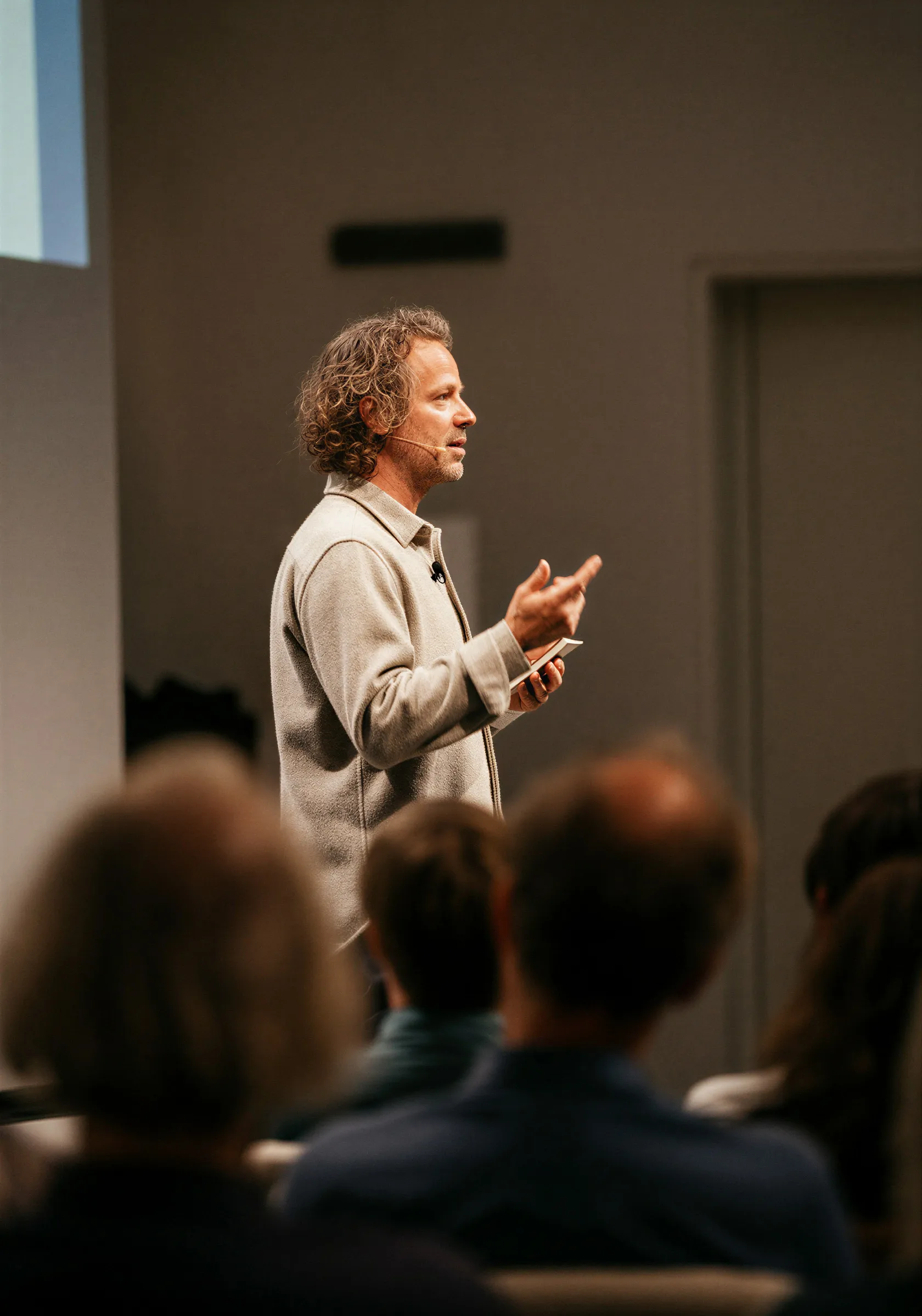 Man with curly hair wearing a beige jacket speaking on stage to an audience while holding a phone.