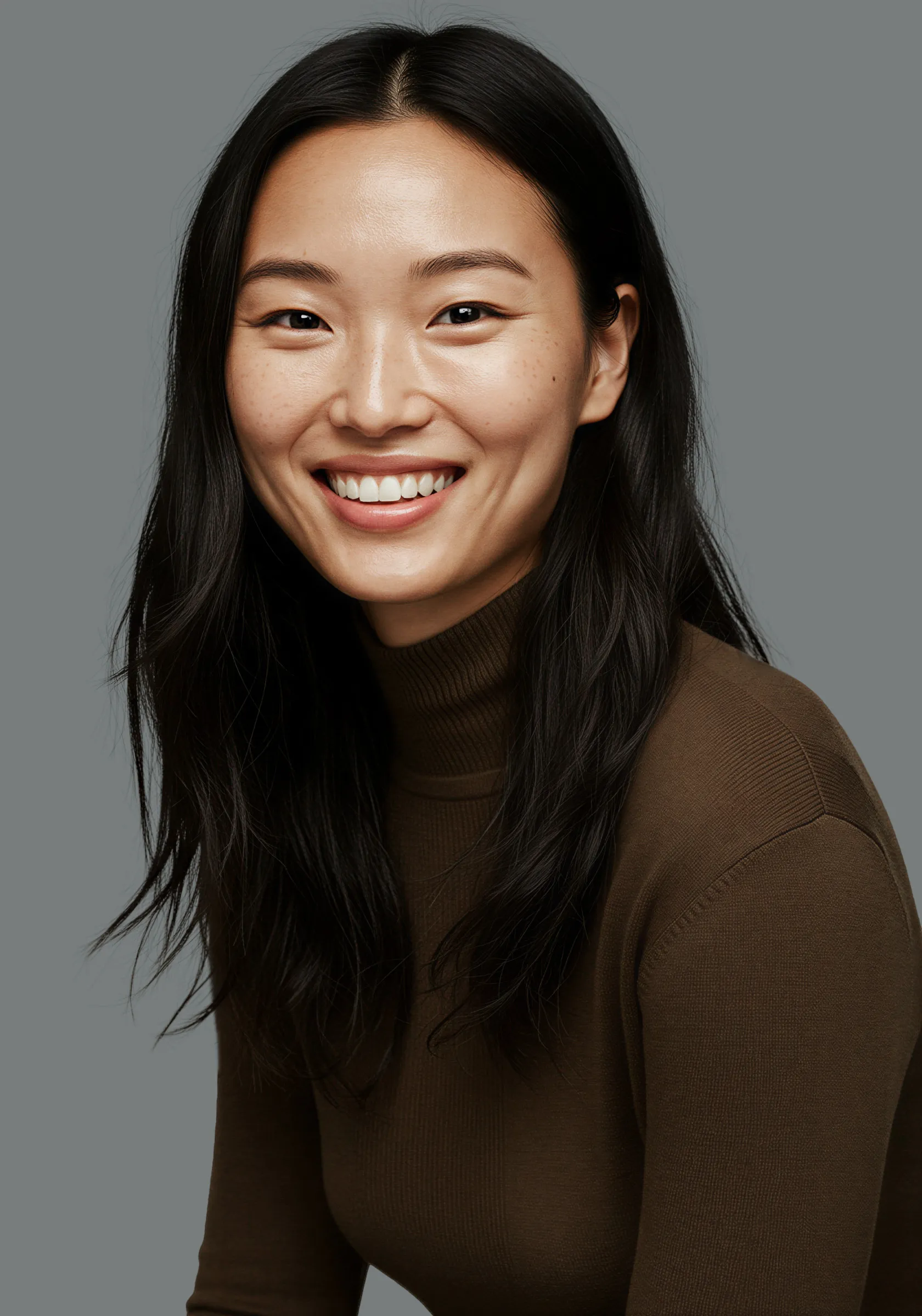 Smiling young Asian woman with long black hair wearing a brown turtleneck sweater against a gray background.