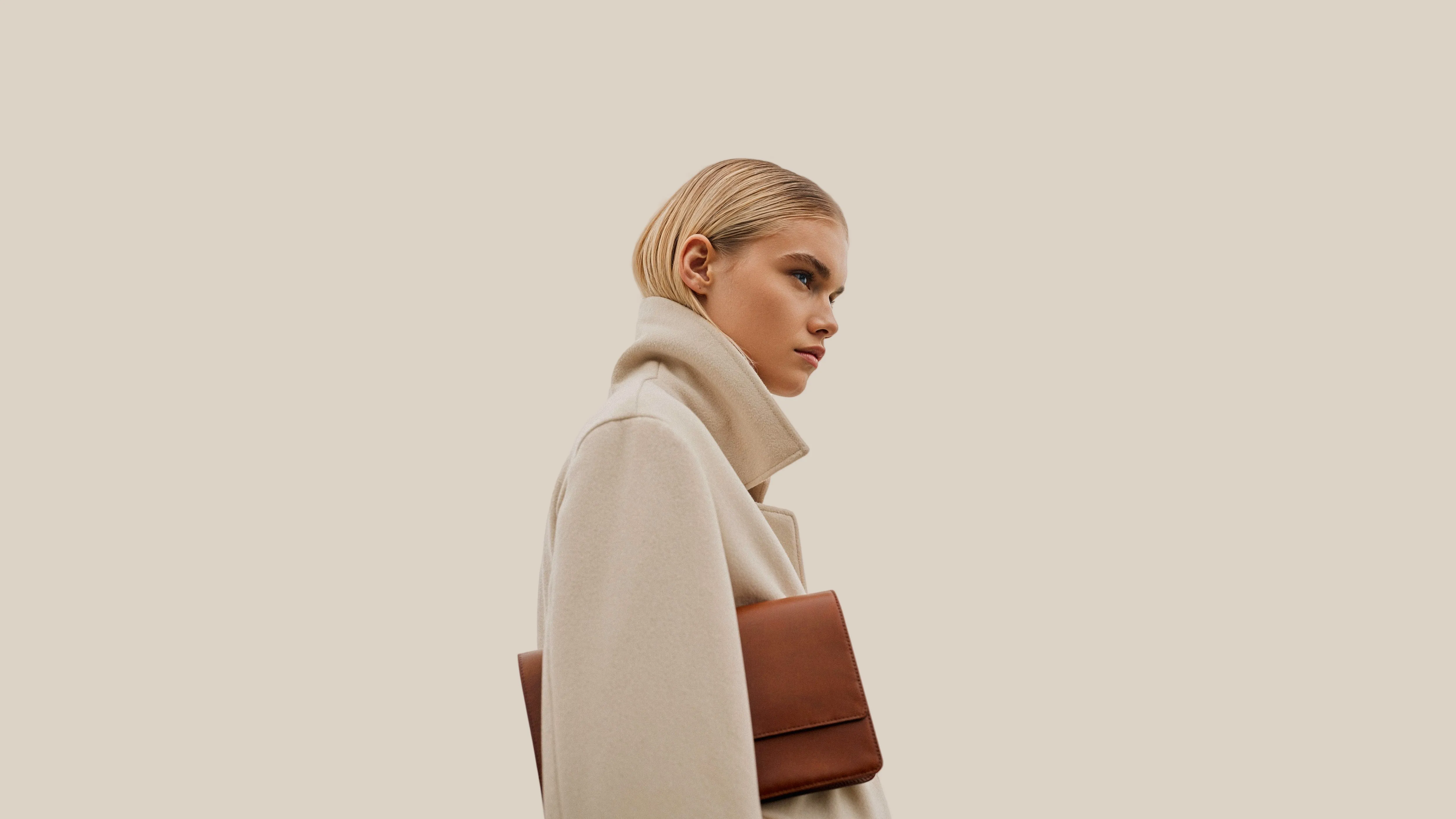 Profile of a woman with short blonde hair wearing a beige coat and holding a brown clutch bag against a neutral background.