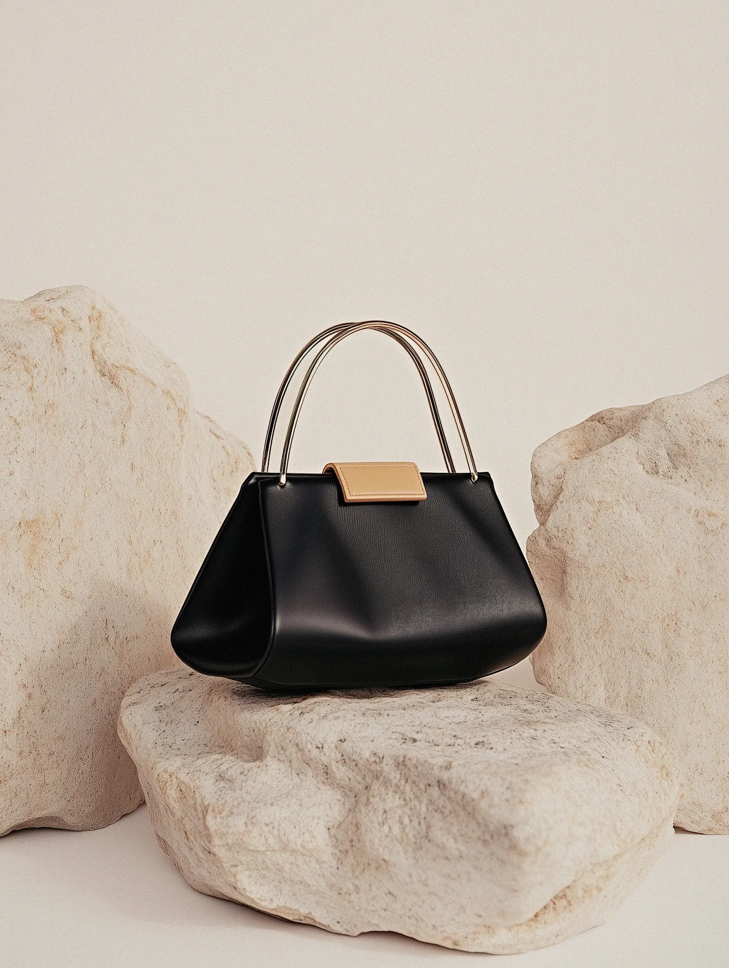 Black leather handbag with gold-tone double handles and beige clasp, placed on textured light stone slabs.