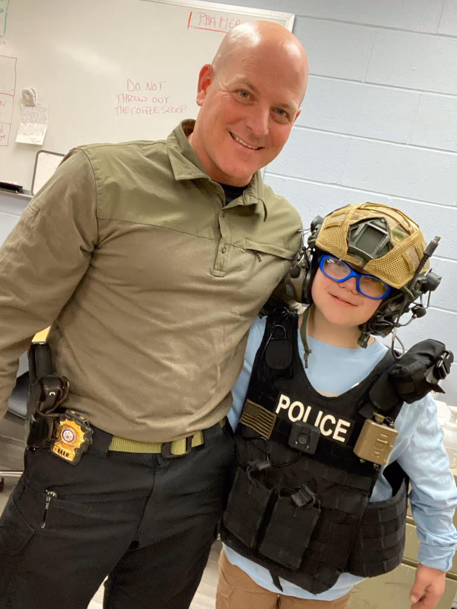 Smiling man in tactical clothing with police badge embraces child wearing police vest, helmet, and glasses in an office.