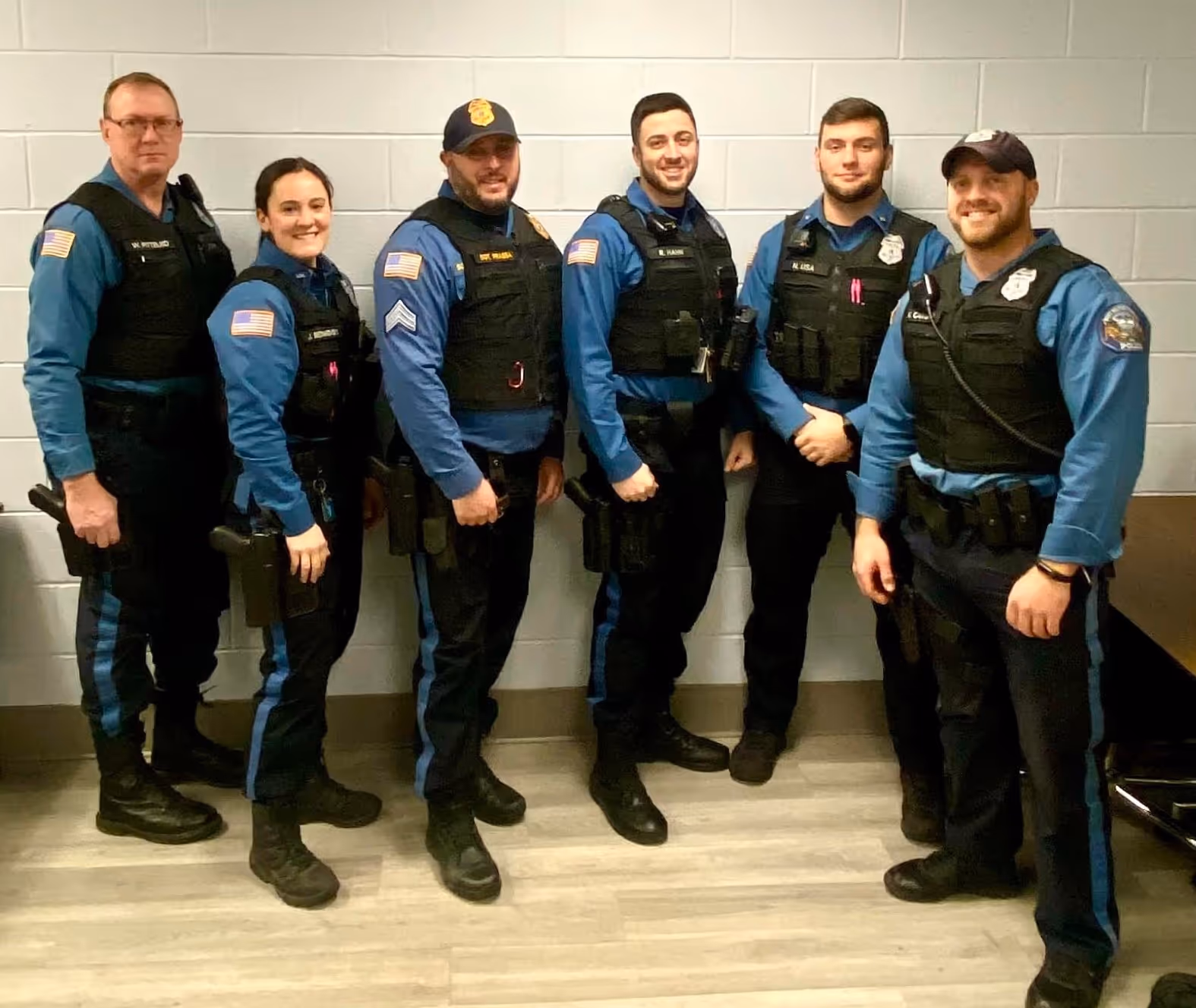 Group of six police officers in blue uniforms and black tactical vests standing indoors against a light gray wall, smiling.