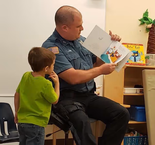 A police officer in uniform reads a children's book aloud to a young boy in a green shirt in a classroom setting.
