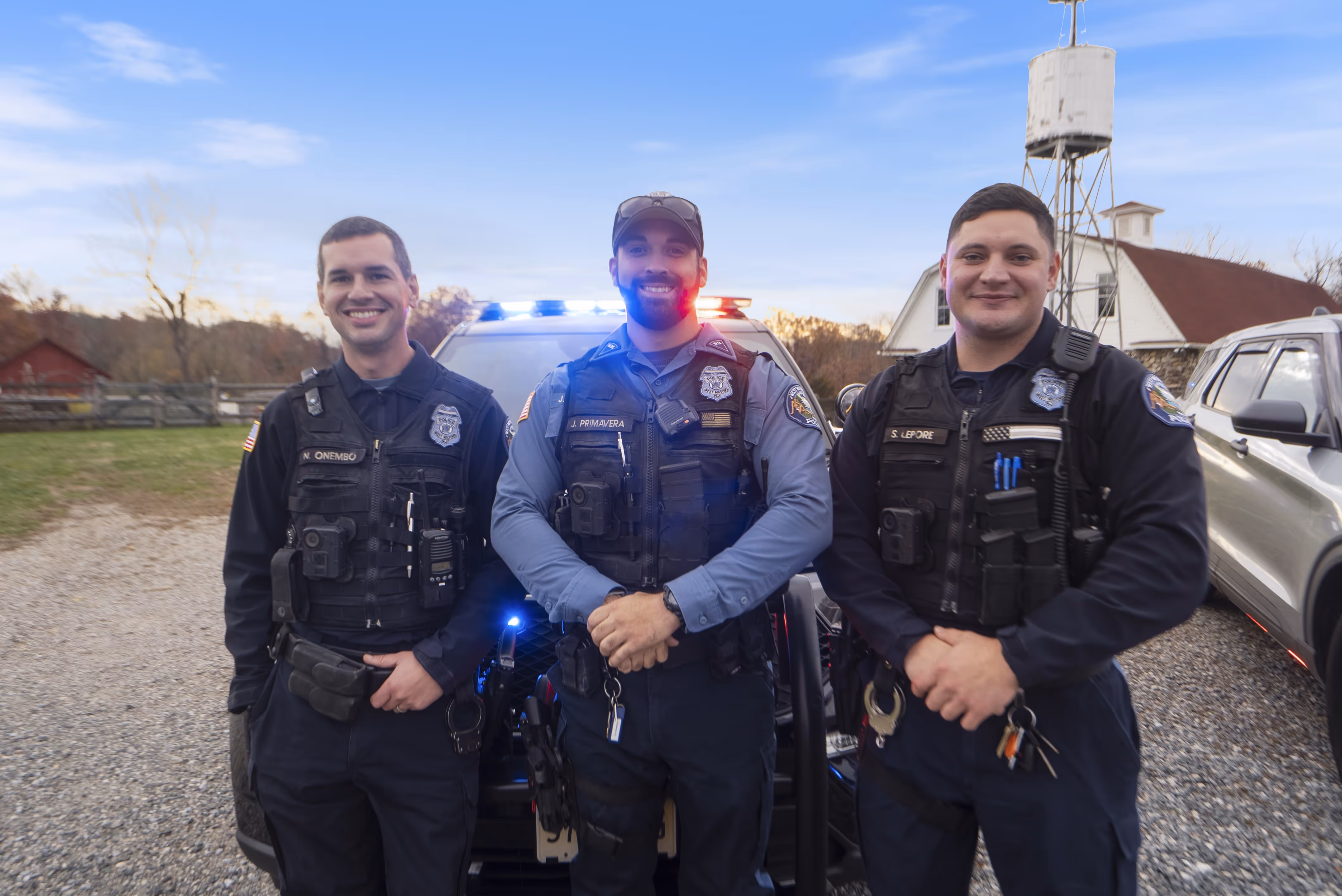 Three smiling police officers standing in front of a police car with flashing lights during sunset.