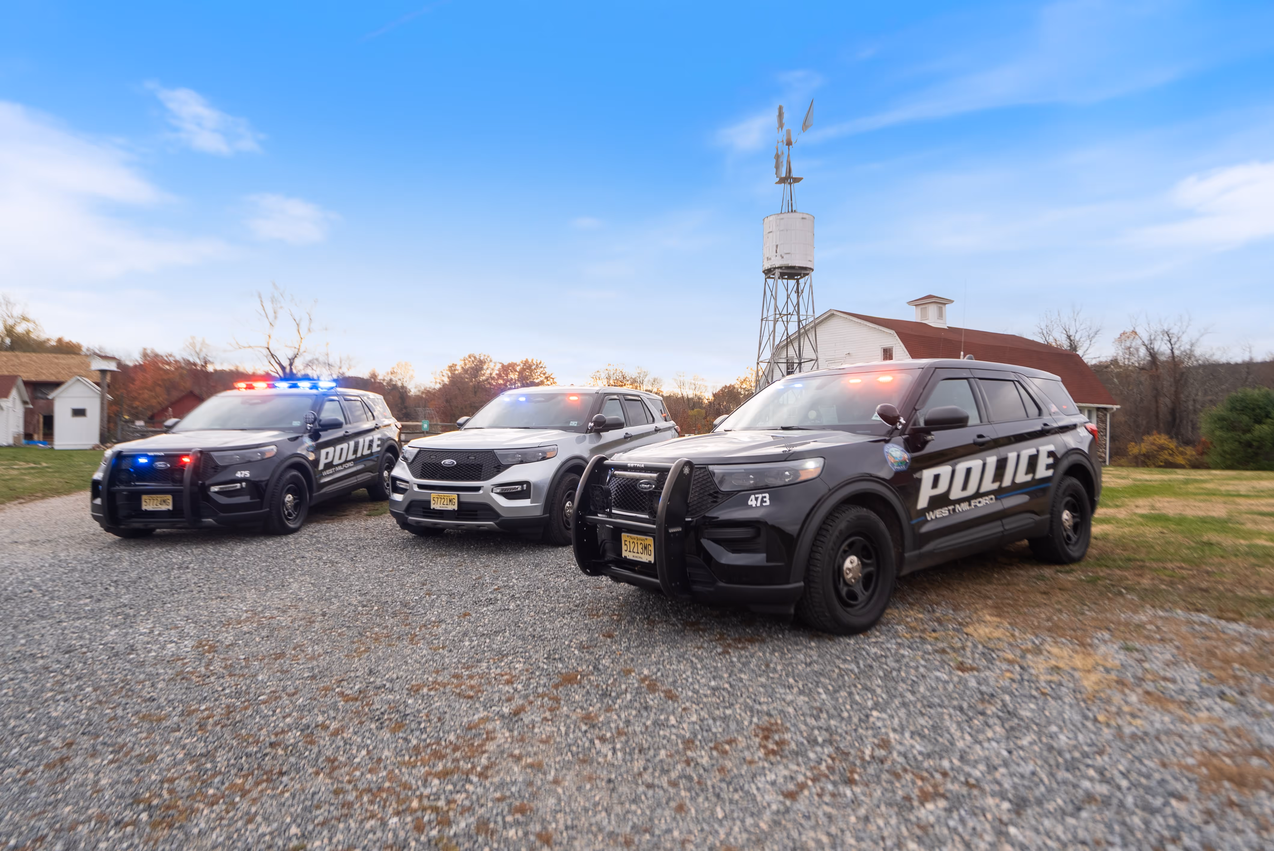 Three West Milford police SUVs with flashing lights parked on a gravel surface near a white building and a windmill under a blue sky.