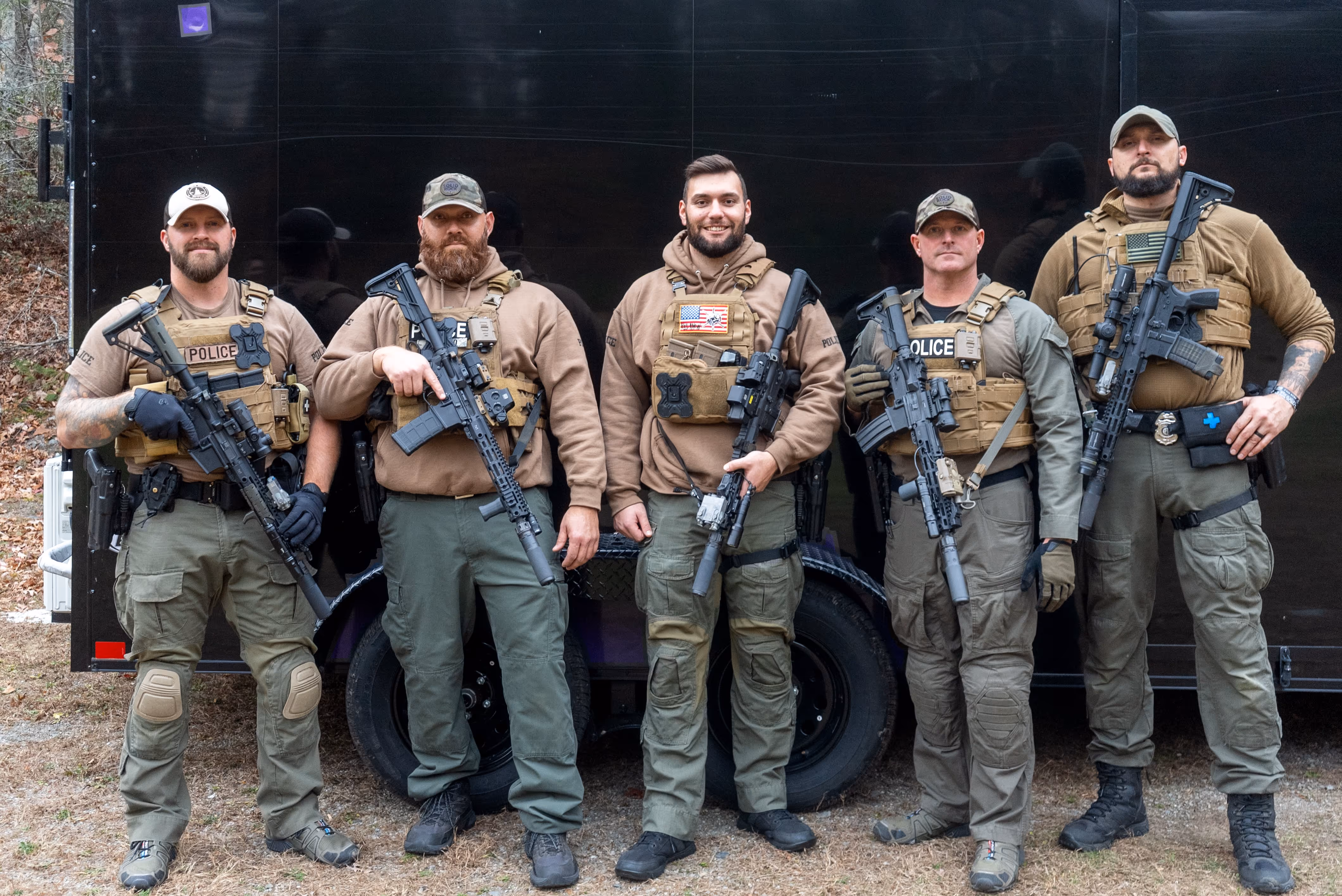 Five armed police officers in tactical gear standing in front of a black vehicle, holding rifles.