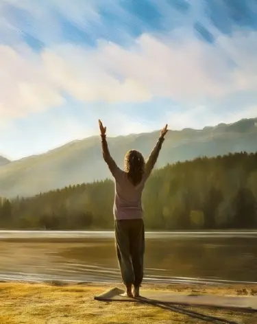 Woman standing at the edge of a lake with both arms raised toward the mountains.