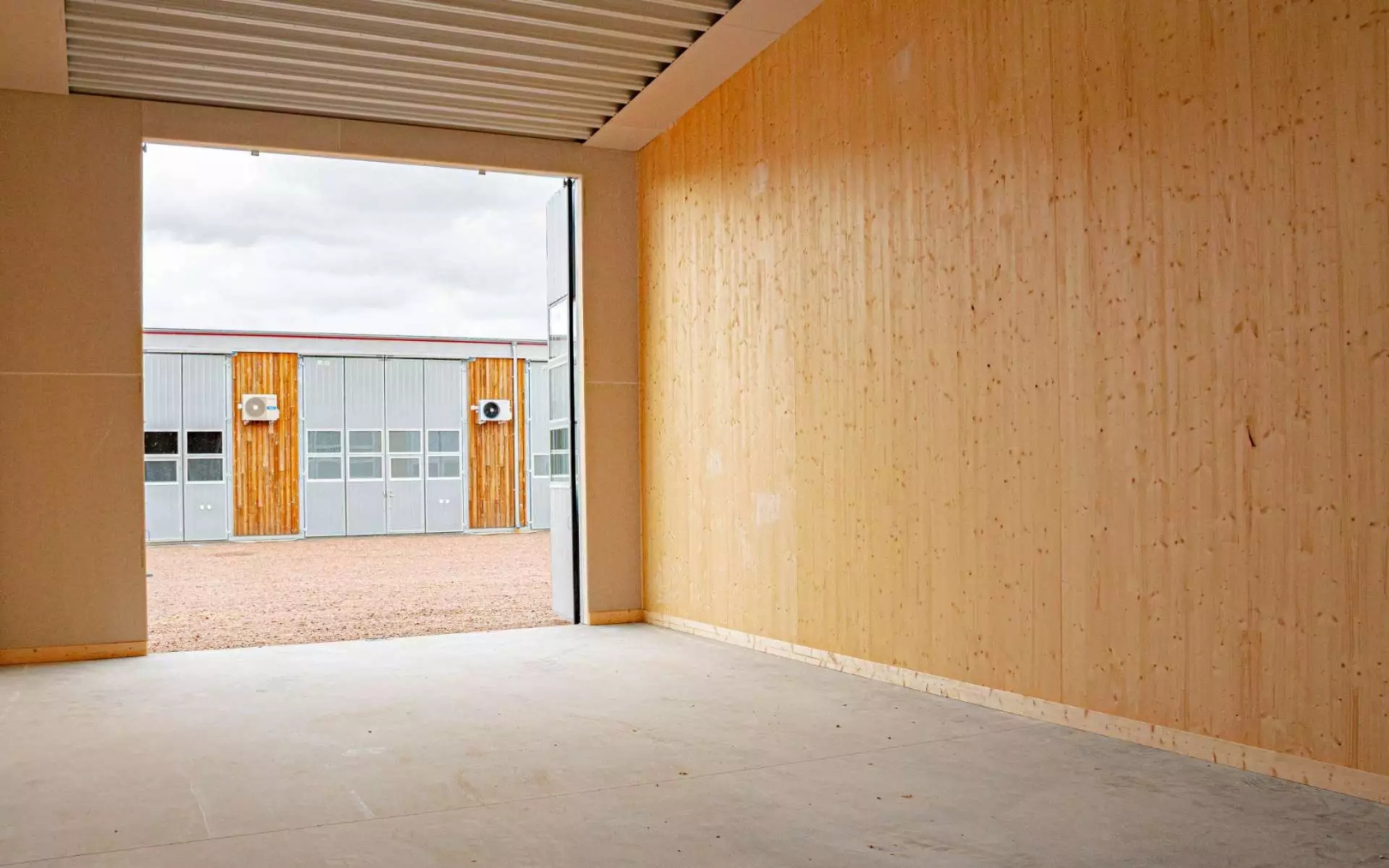 Tom lagerlokal med betonggolv, träpaneler på väggarna och ett öppet port med utsikt mot en grusgård och en byggnad med metall- och träytor.