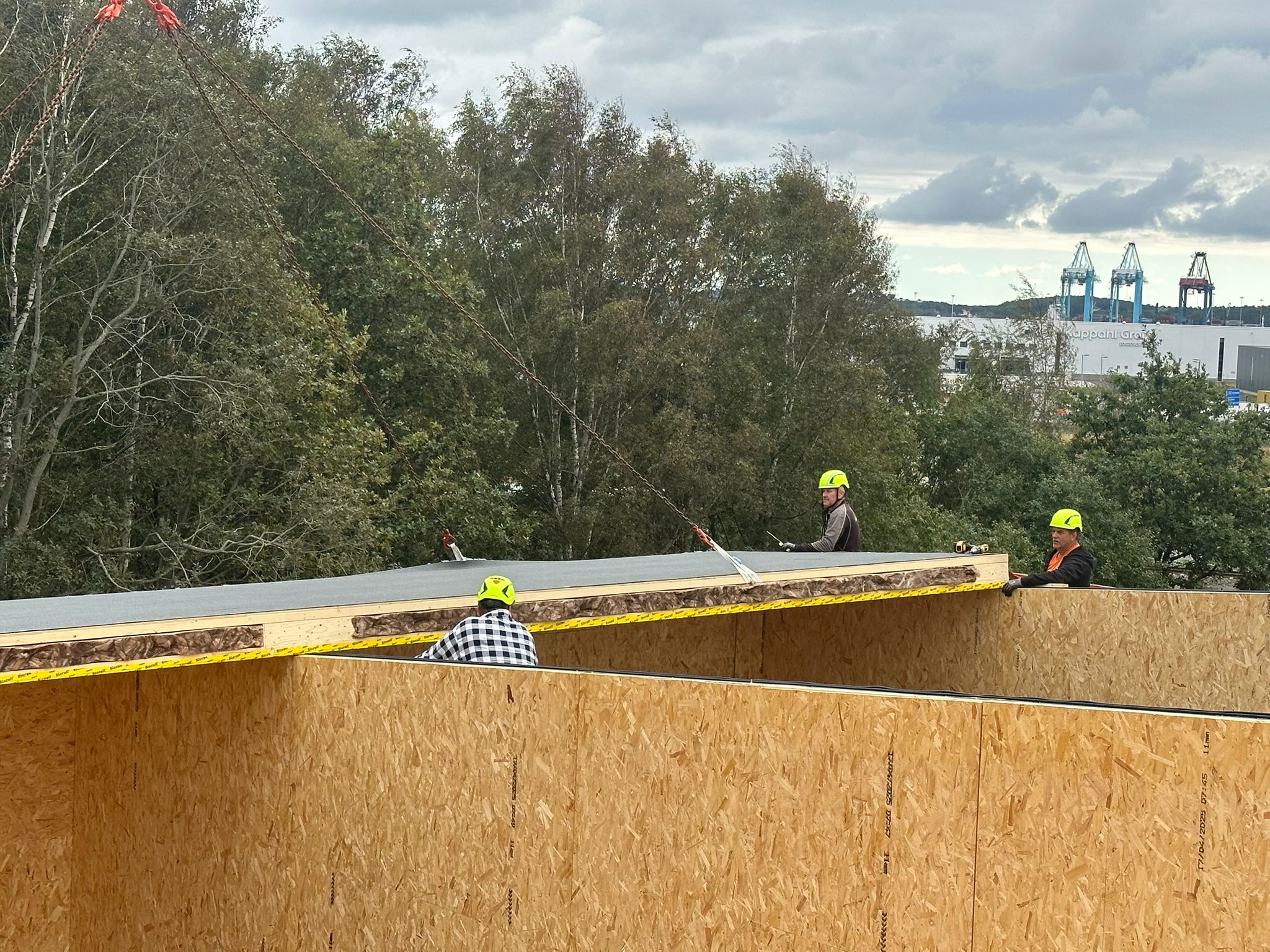 Byggarbetare med gula hjälmar lyfter och placerar en stor takpanel på en träkonstruktion under ett molnigt himmel, med träd och industriområde i bakgrunden.