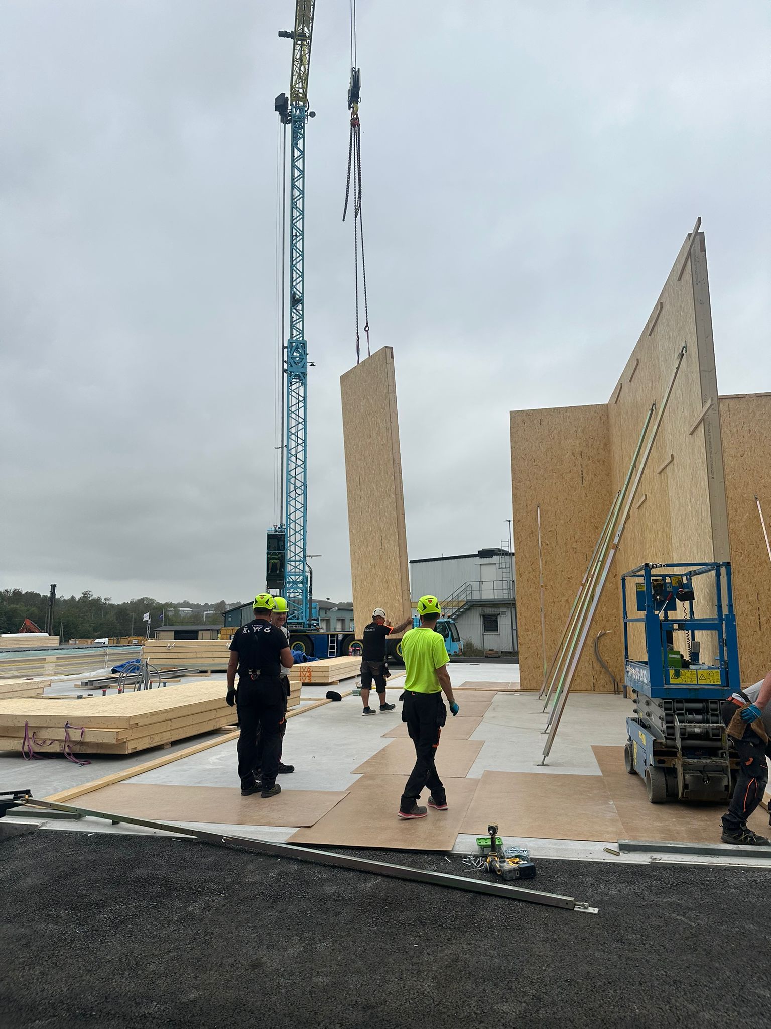 Byggarbetare som monterar stora träpaneler på en byggarbetsplats med en kran under molnig himmel.