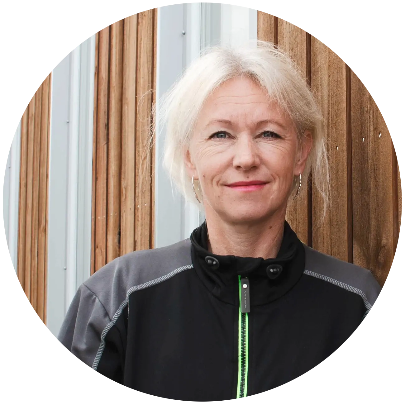 Porträtt av en kvinna med blont hår och svart jacka framför en trävägg.