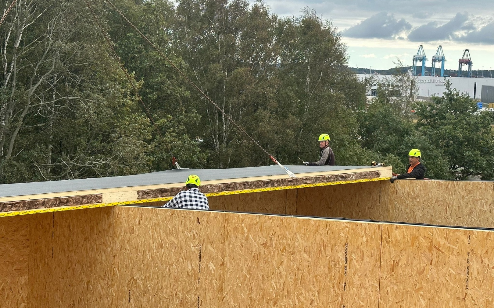 Tre byggnadsarbetare med gula hjälmar lyfter ett stort träelement på en byggarbetsplats med träd i bakgrunden.