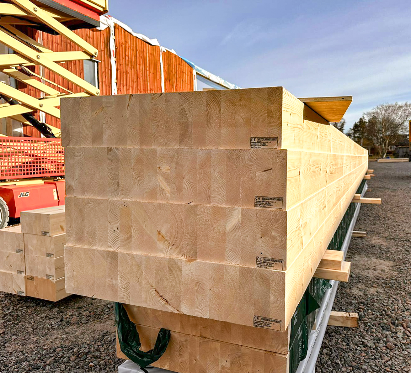 Staplad limträbalkar på byggarbetsplats med röd byggnadsställning och hus i bakgrunden.