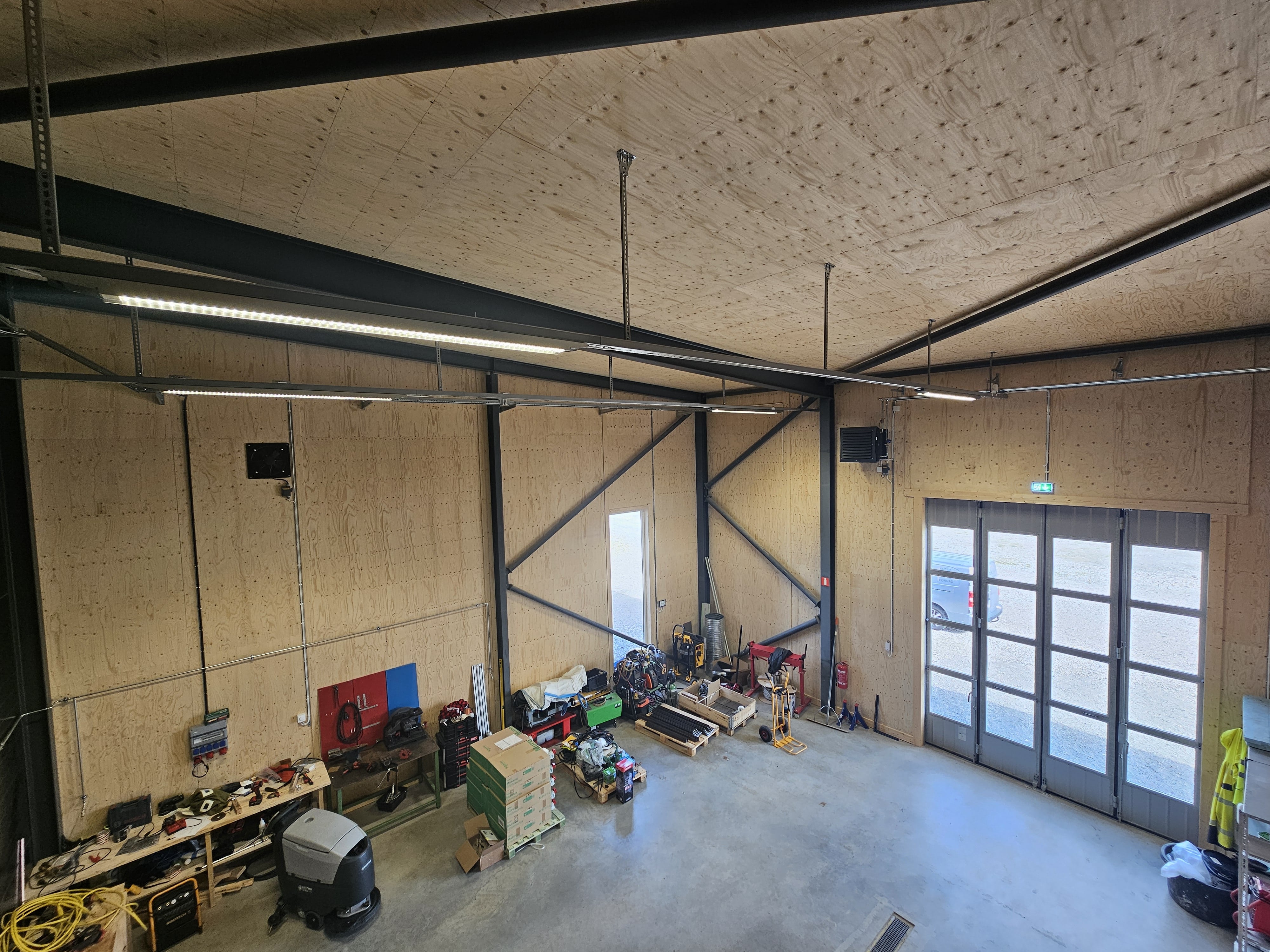 Stor verkstad med plywoodväggar, arbetsbord, verktyg, kartonger och stora fönster med dörrar ut mot en uppfart.