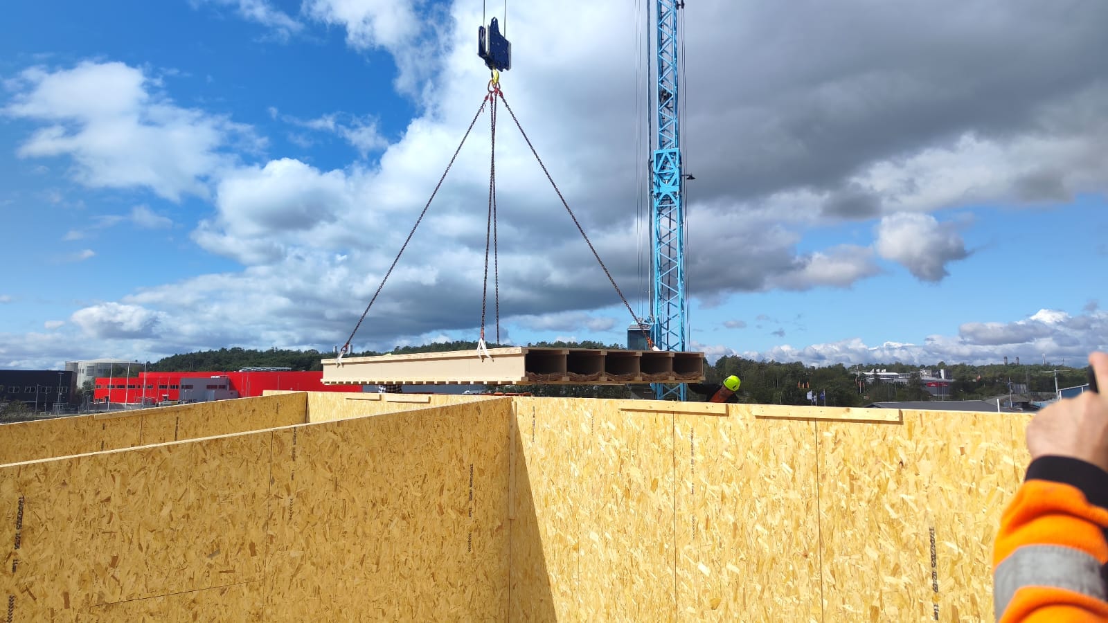 Byggarbetsplats med kran som lyfter en stor träpanel över träväggar under en blå himmel med moln.