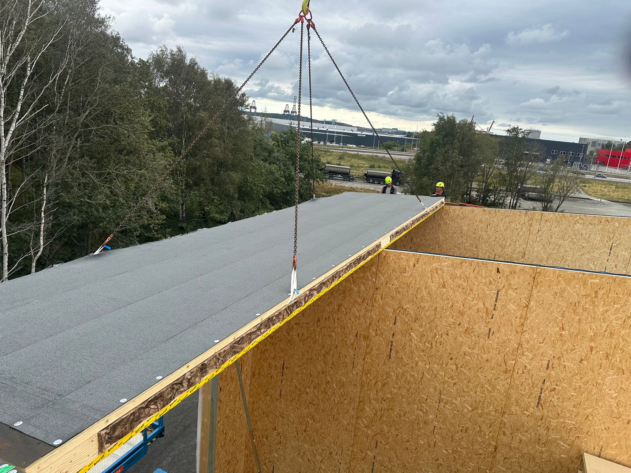 Byggnadsarbetare med hjälmar installerar ett tak av träpaneler med hjälp av en kran på en byggarbetsplats.