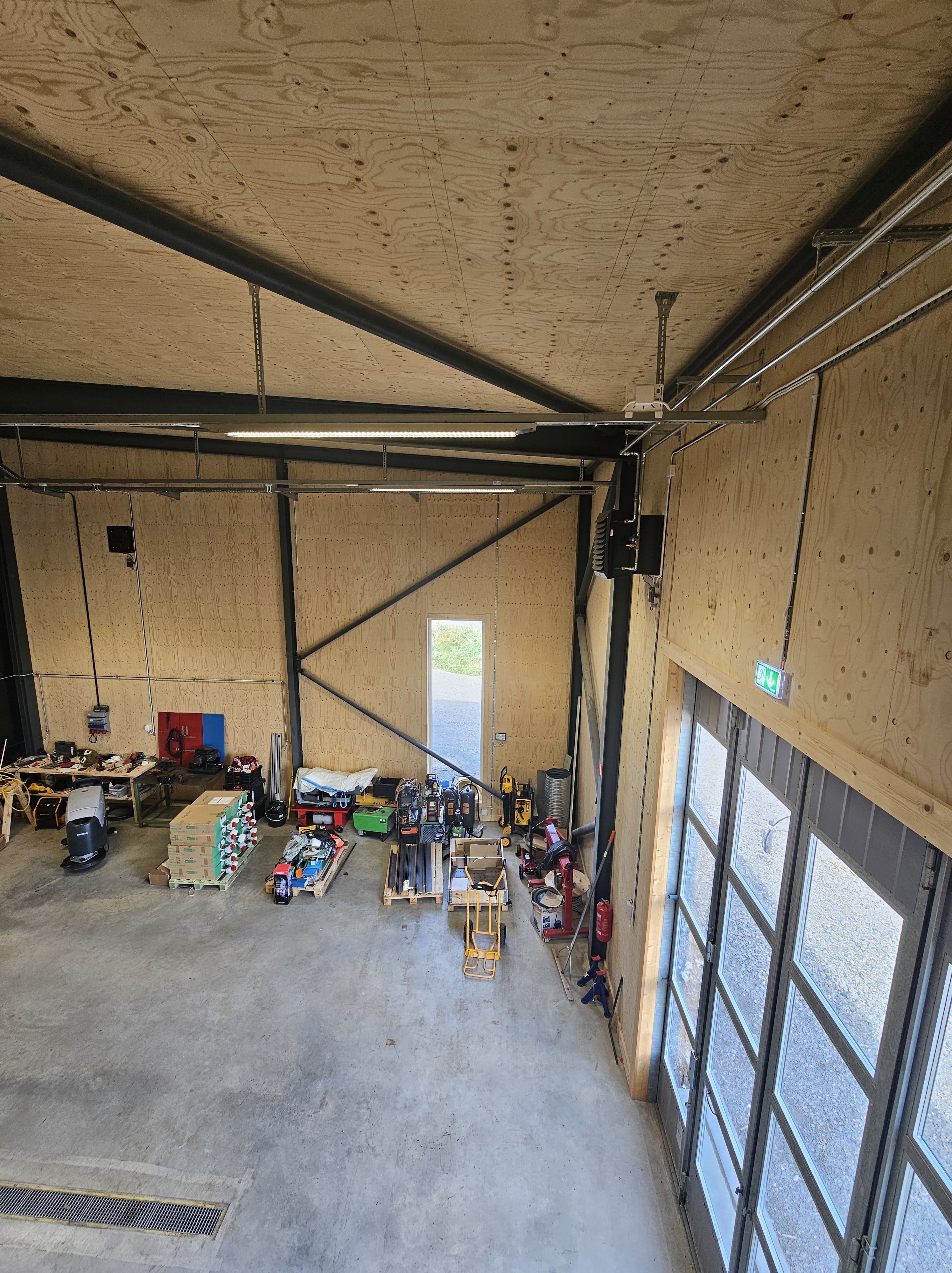 Inre vy av en verkstad med verktyg, maskiner och förvaringspallar längs en vägg av träpaneler.