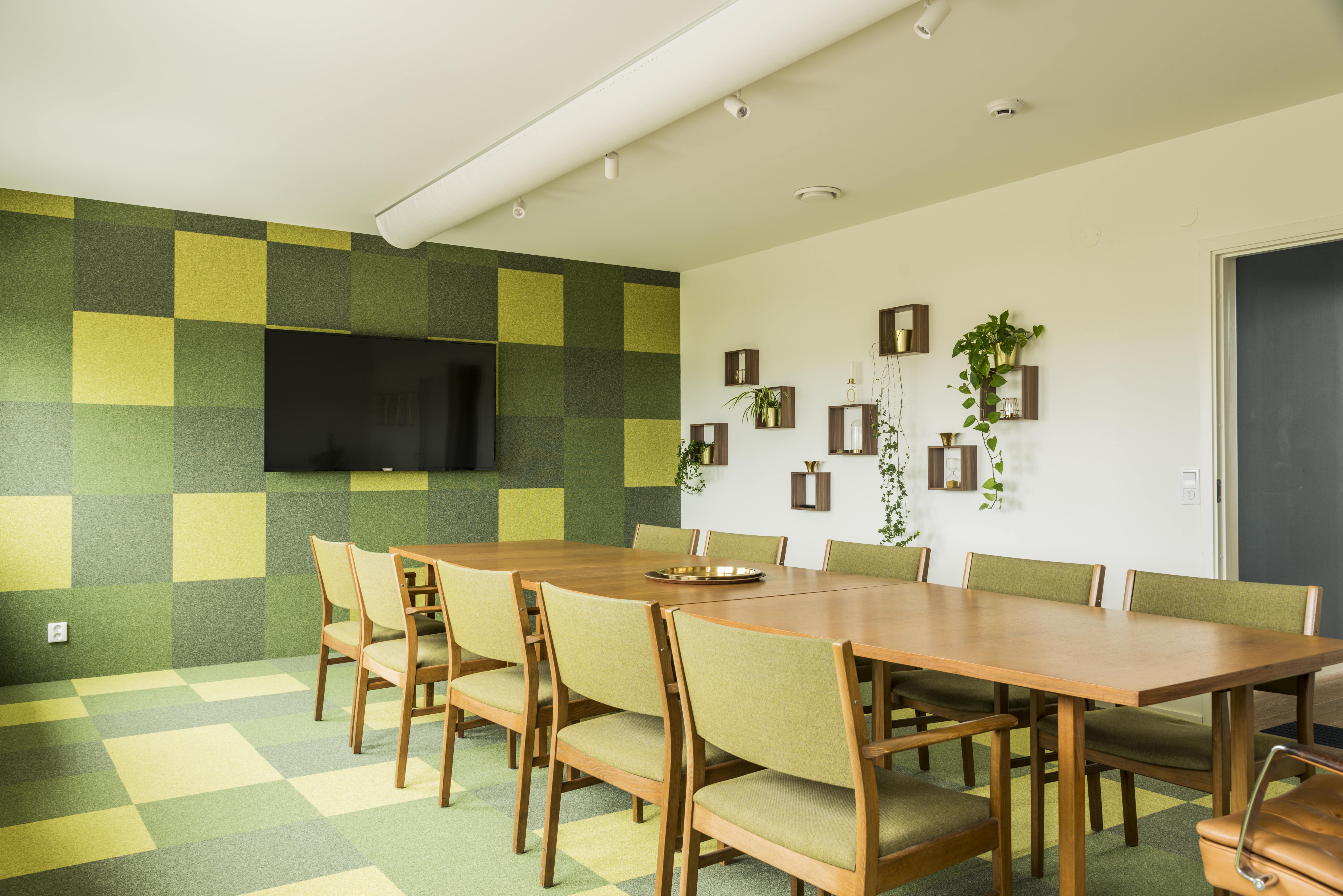 Konferensrum med långt träbord och gröna stolar, med en vägg täckt av gröna rutmönster och växtdekorationer på vita vägghyllor.