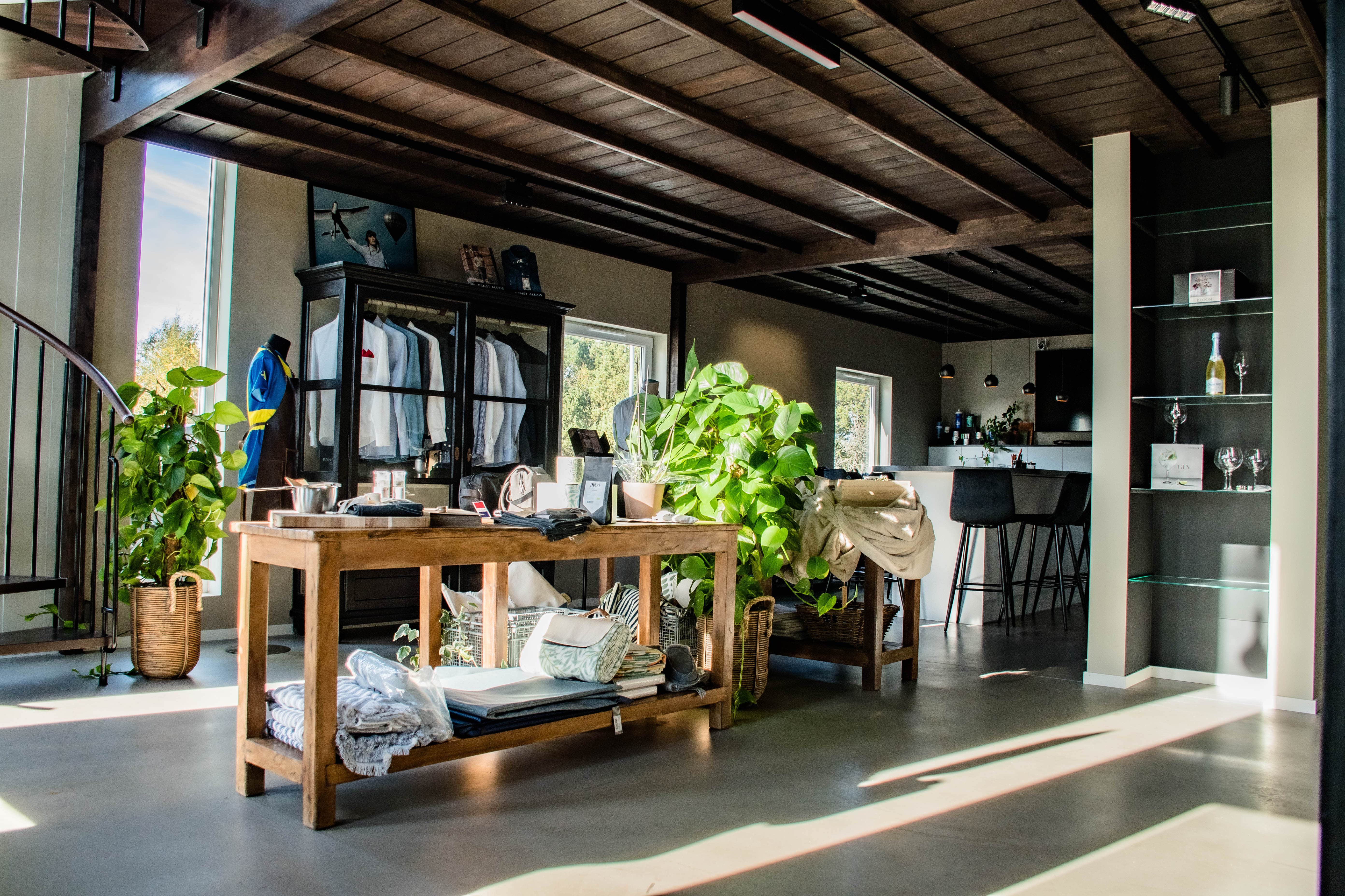 Ljus och modern butiksmiljö med kläder på galgar, trämöbler, gröna växter och barstolar vid en bardisk.