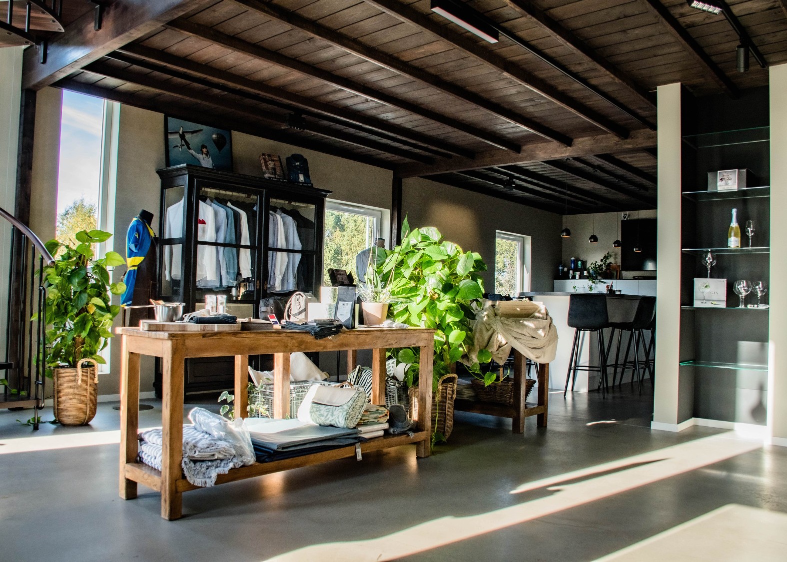 Ett modernt rum med ett träbord fyllt med vikta kläder och växter, en garderob med vita skjortor, samt en köksö med barstolar i bakgrunden.