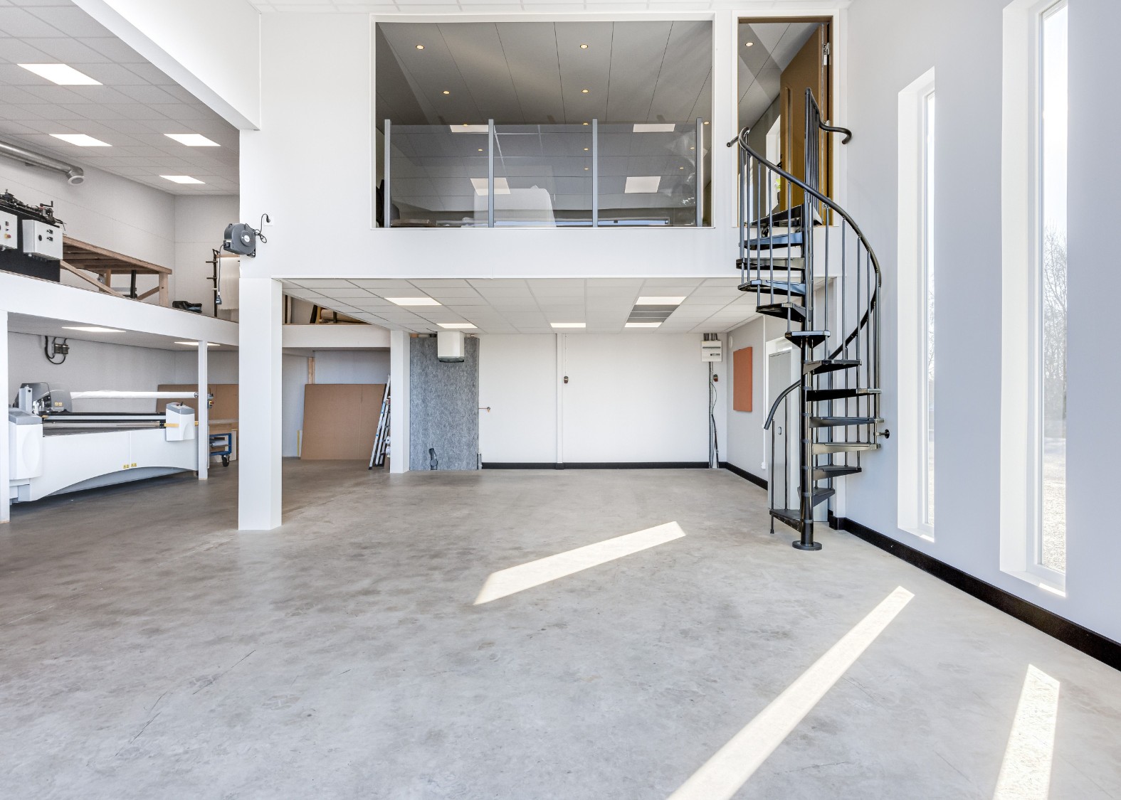 Ljust, tomt industrilokal med betonggolv, spiraltrappa och stora smala fönster som släpper in dagsljus.