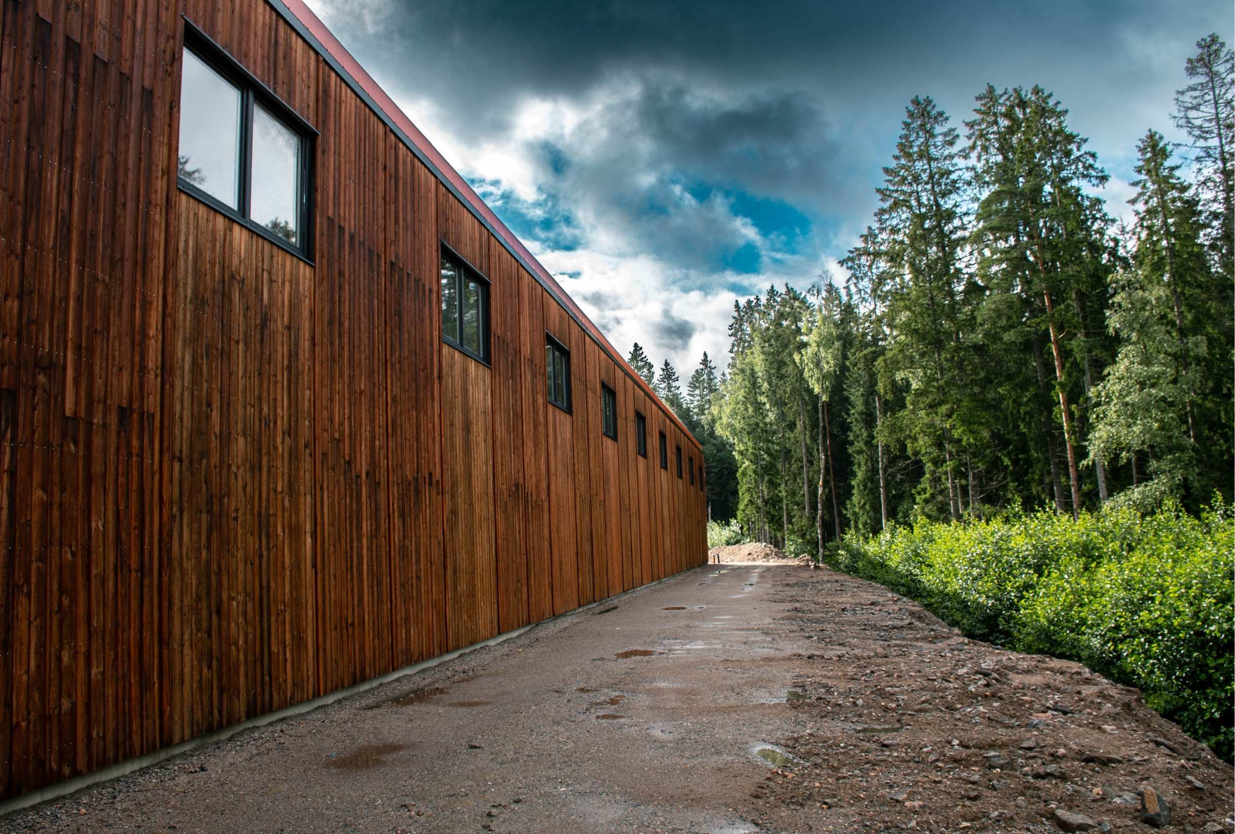 Lång byggnad med träpanel och små fönster intill skog och en grusväg under en molnig himmel.