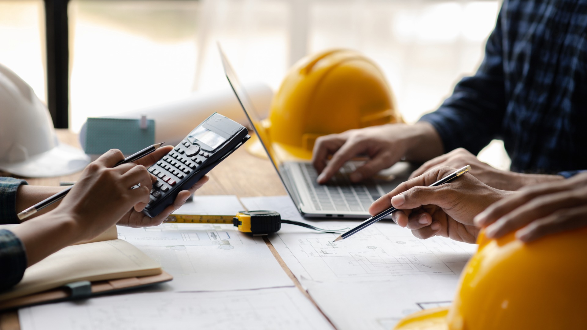 Två personer granskar tekniska ritningar, en använder miniräknare och en annan använder laptop, med gula skyddshjälmar på bordet.