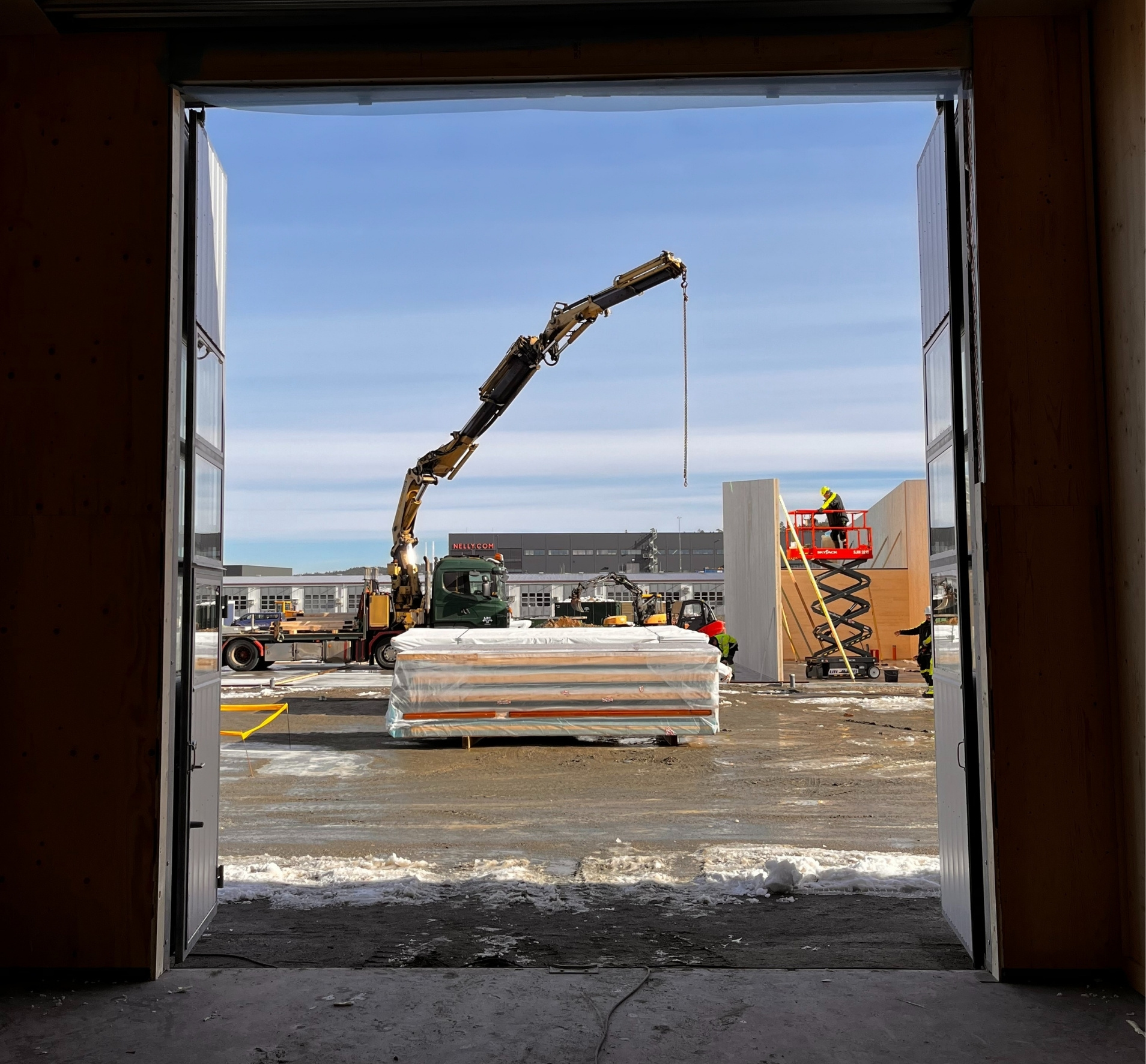 Byggarbetsplats sedd genom en öppen port med en kranbil och arbetare på en lyftplatta som monterar väggar.