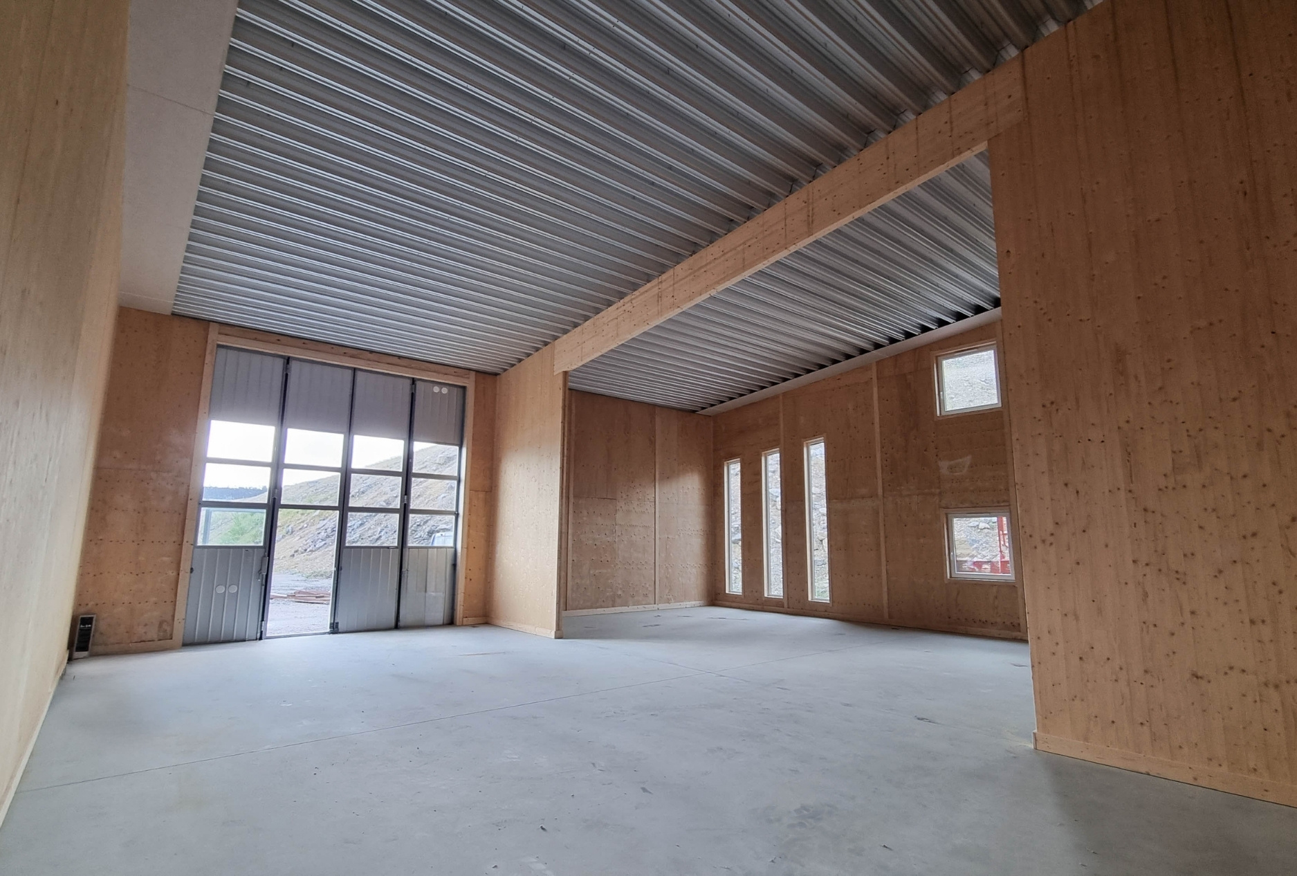 Stor tom industrilokal med träväggar, betonggolv, metalltak och stora fönster samt dörr som vetter mot utsida.