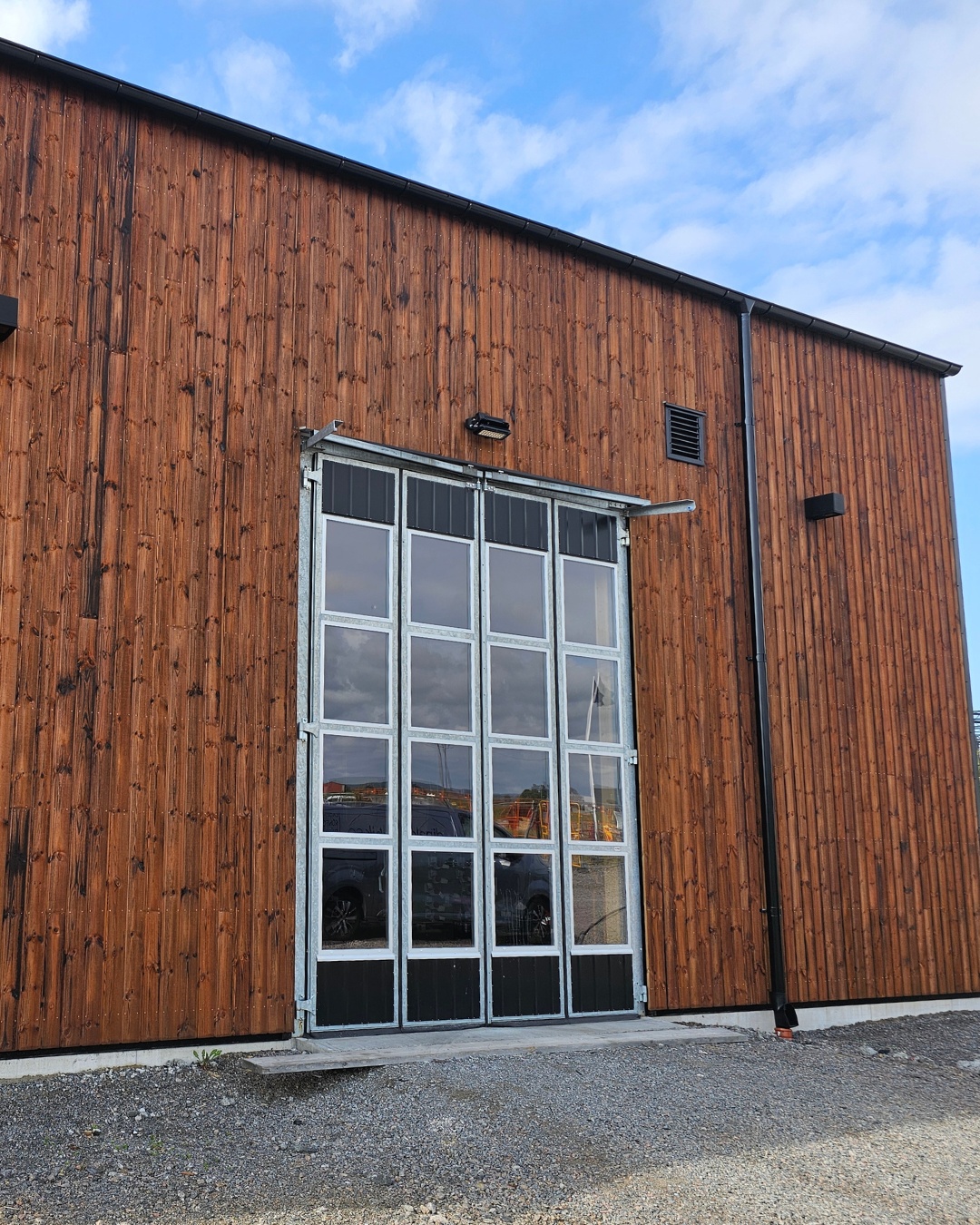Stor träbyggnad med stora glasade industridörrar och en grusbelagd uppfart under en blå himmel.