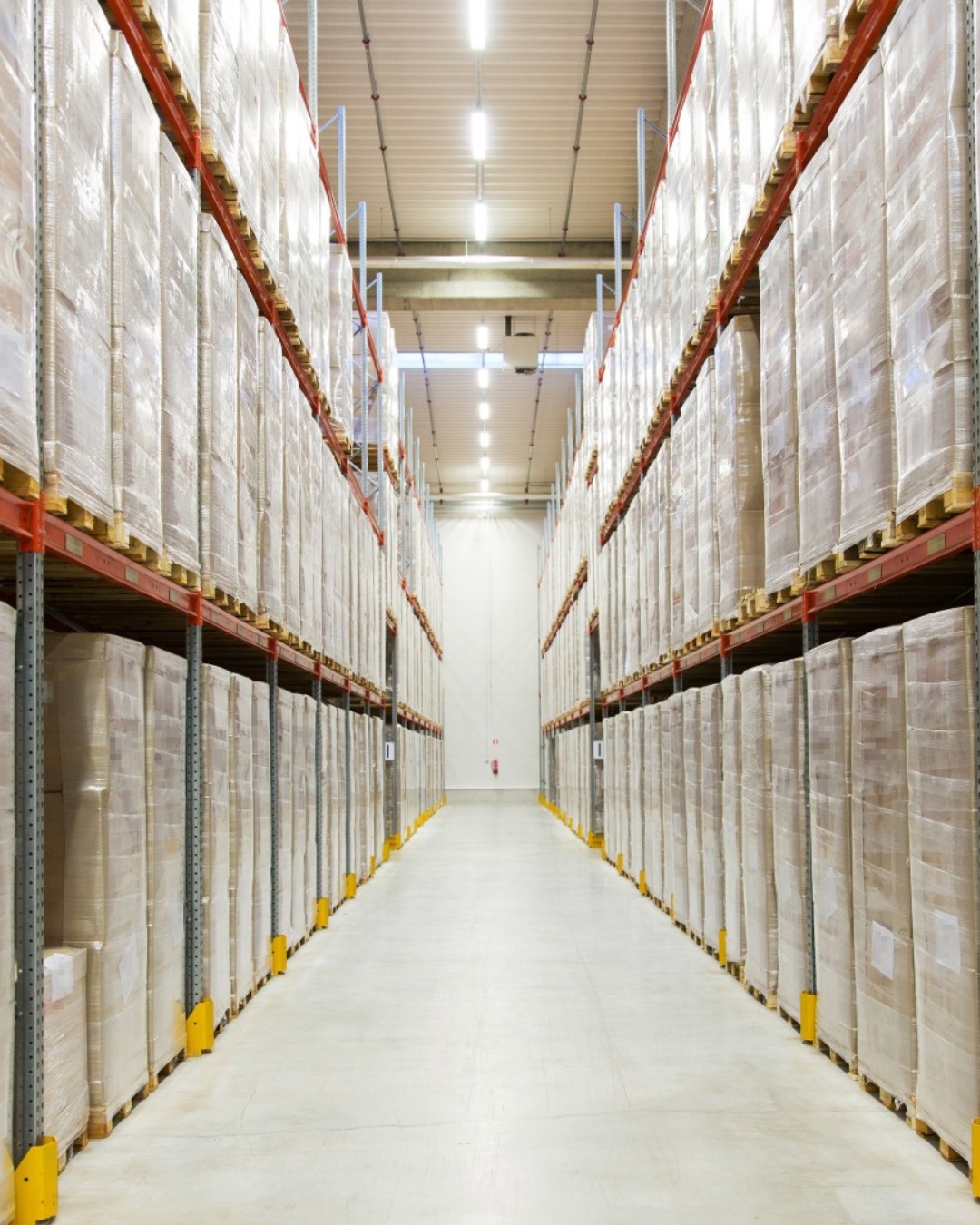 Lång gång i ett lager med höga hyllor fyllda med staplade och plastade kartonger.