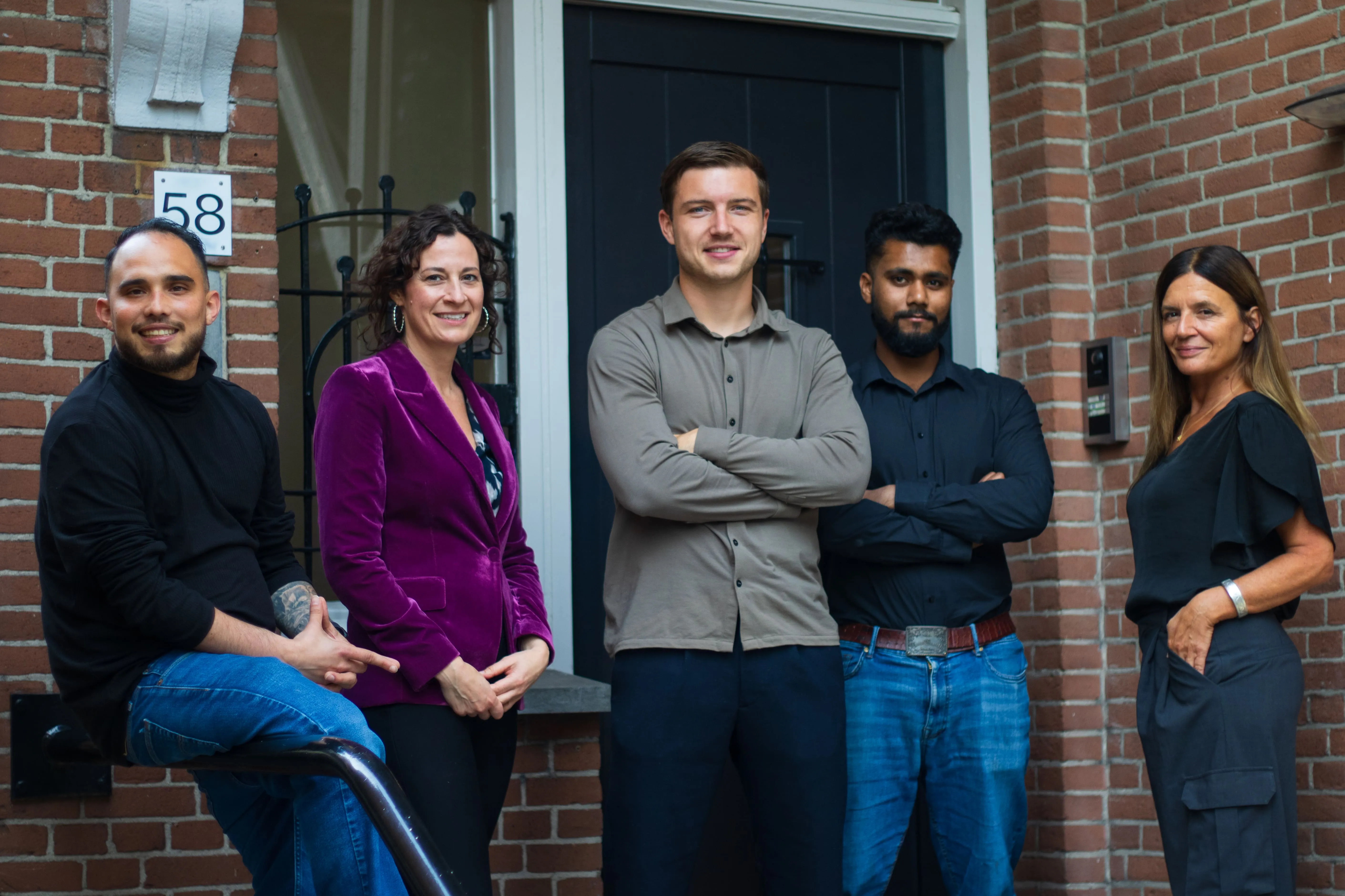 Group of five people posing in front of a brick building entrance with a black door, smiling and standing confidently.