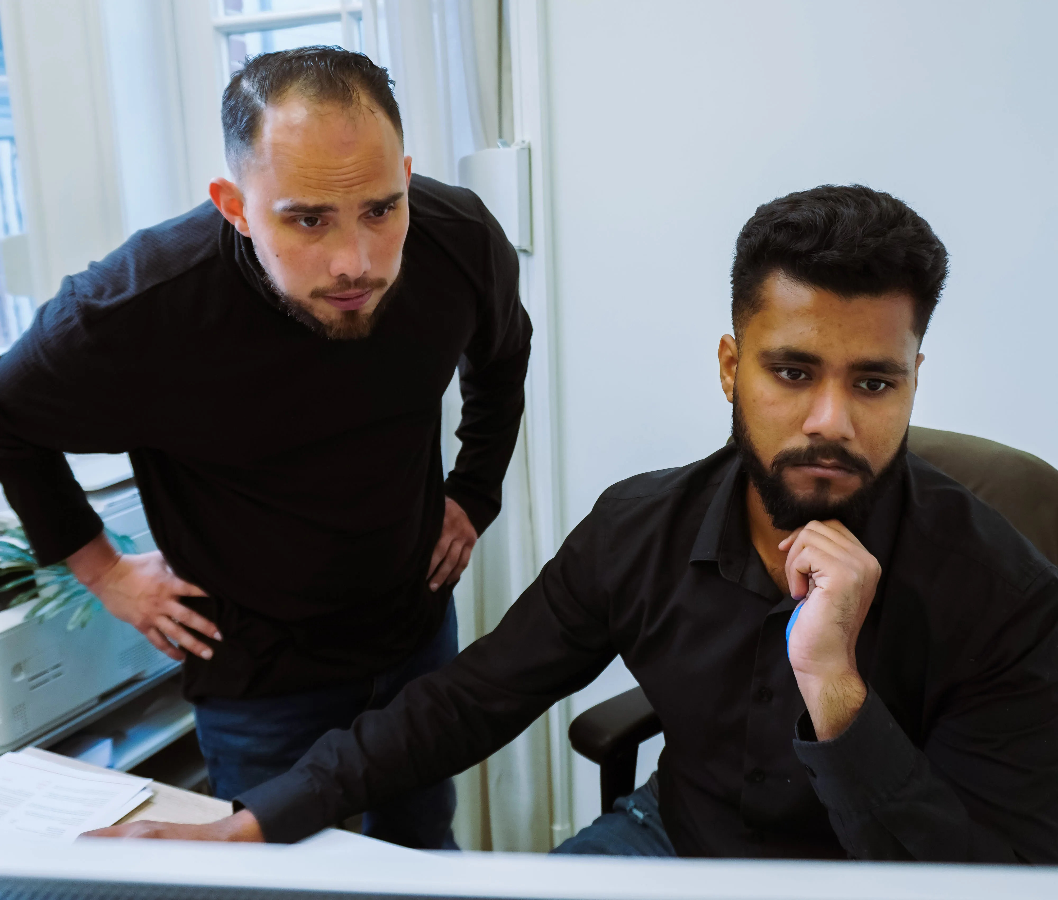 Two men in black shirts focused on a computer screen; one seated with hand on chin and the other standing with hands on hips.