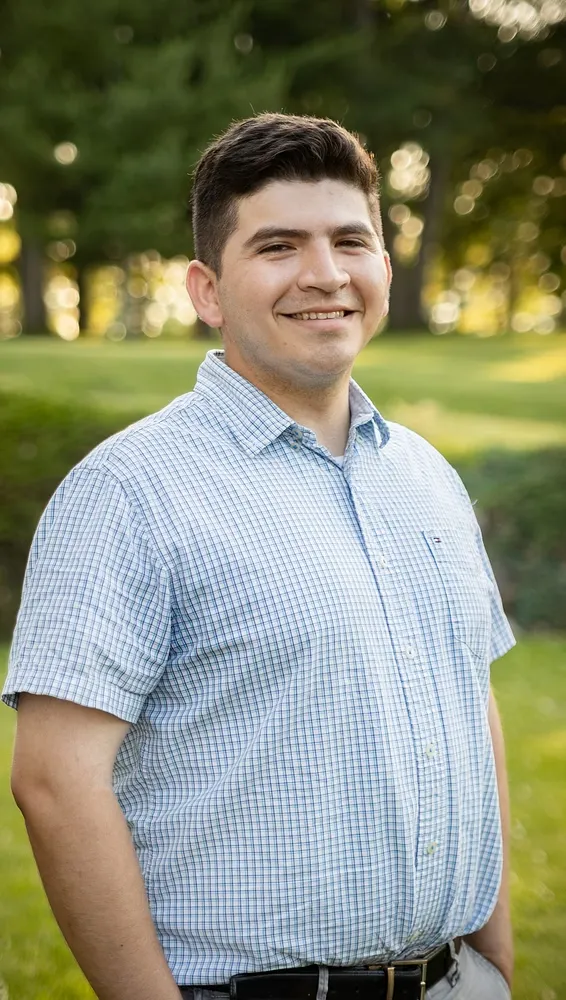 Smiling man in a short-sleeve checkered shirt standing outdoors with trees and grass in the background.