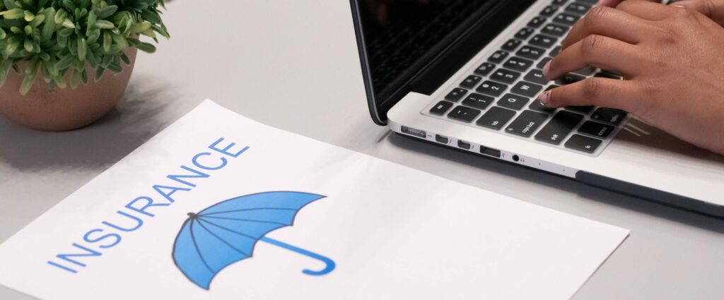 Someone is working on their laptop. Beside the laptop is a piece of paper featuring the word “Insurance” in blue text over the image of a blue umbrella
