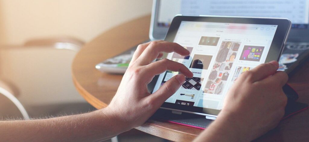 A close-up of a woman’s hands as she works on a tablet. A laptop is in the background.