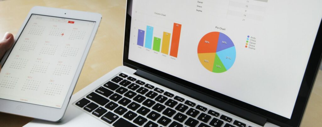 A photo of a laptop displaying a pie chart and graph on its screen. Someone holds a tablet beside it. 