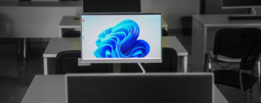 A photo of an empty computer classroom. A single monitor is on featuring an abstract blue image.
