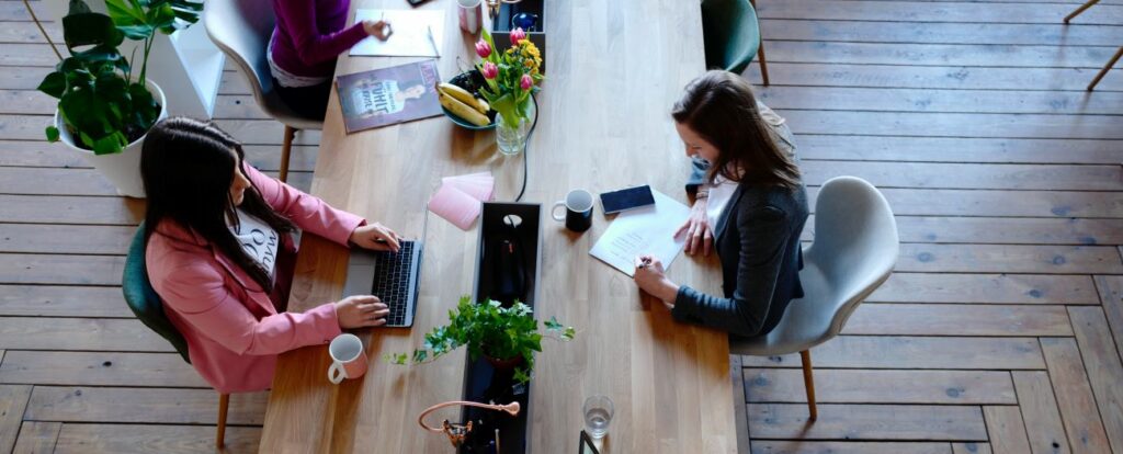 An overhead photograph of four women in business attire sitting at a long wooden table. Two women are working on laptops, and the others are writing on paper.