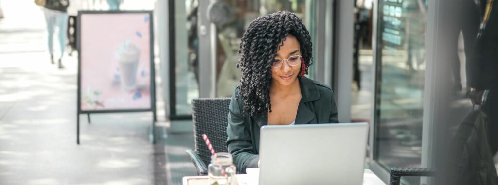 A photograph of a woman cheerfully working on her laptop outside a café. 