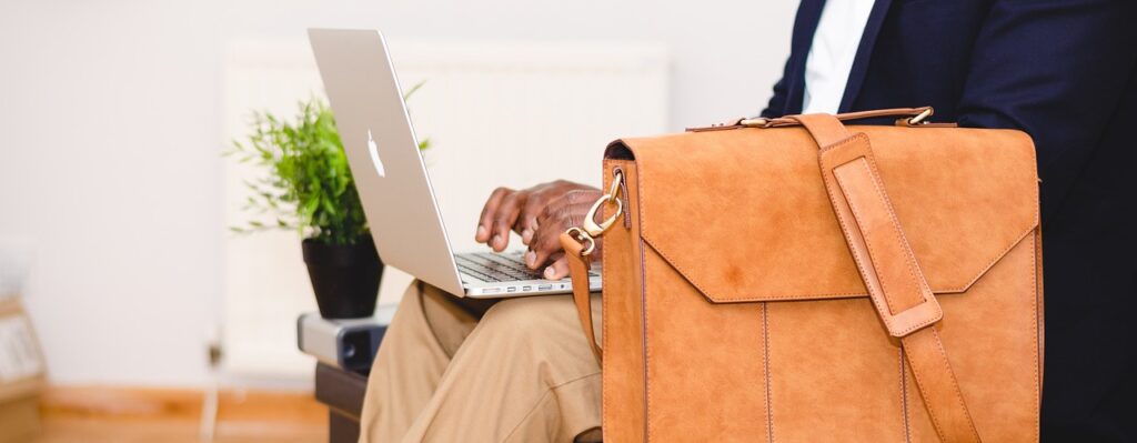 A photograph of a man typing on his laptop and sitting on a leather bench. A light leather briefcase sits beside him on the bench.