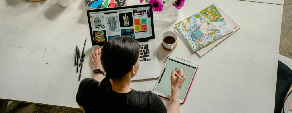A overhead shot of a man at a white desk. He is creating a graphic design both on his laptop and on a tablet.