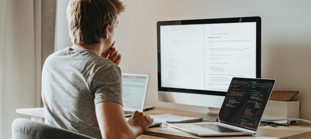 A man looks at his monitor. He has two laptops open on his desk.