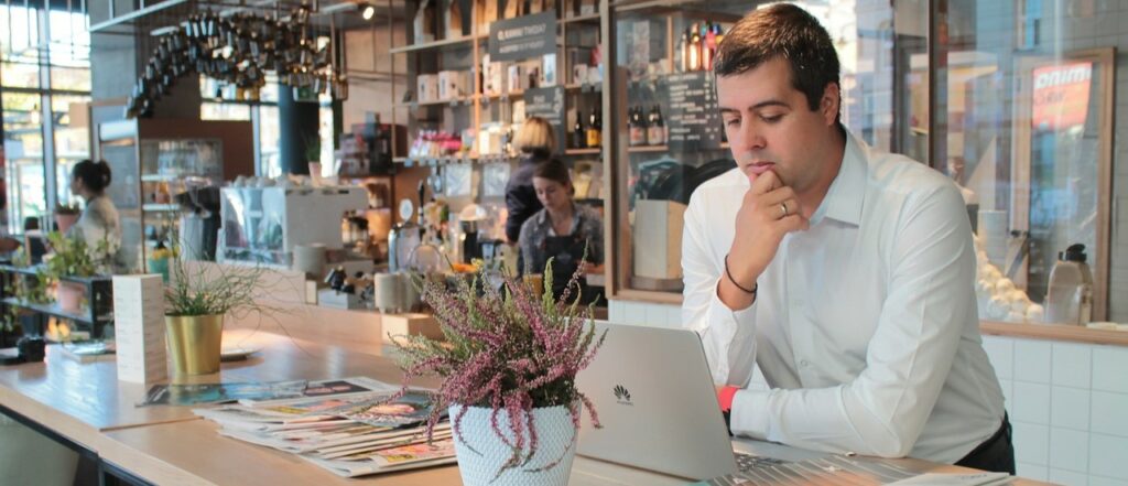 A photograph of a man working on a laptop at a cafe counter.
