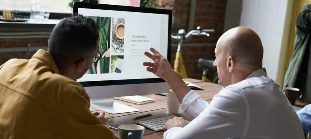 A photograph of two men discussing a website layout displayed on a computer monitor.