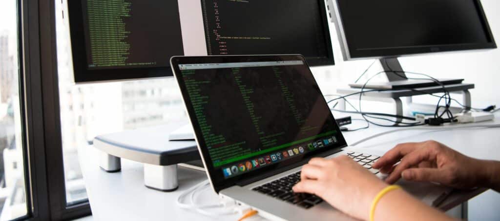 A photo of a man typing on a laptop. His laptop screen and three large monitors behind it all display green coding text.
