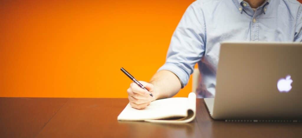 A photograph of a man working at a laptop and taking notes in a notebook.