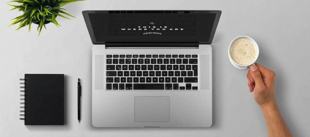 A overhead photograph of a laptop, a notebook, and someone holding a cup of coffee.