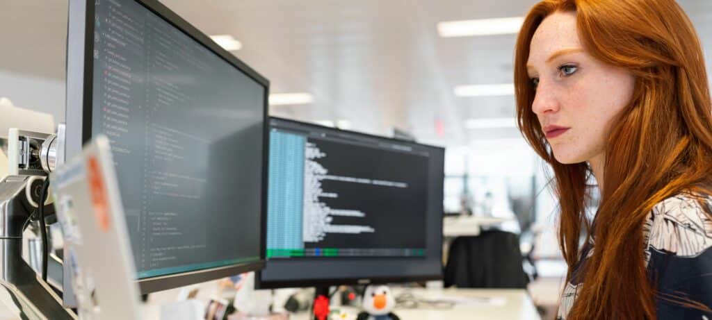 A photograph of a woman working on two computer monitors and a laptop.